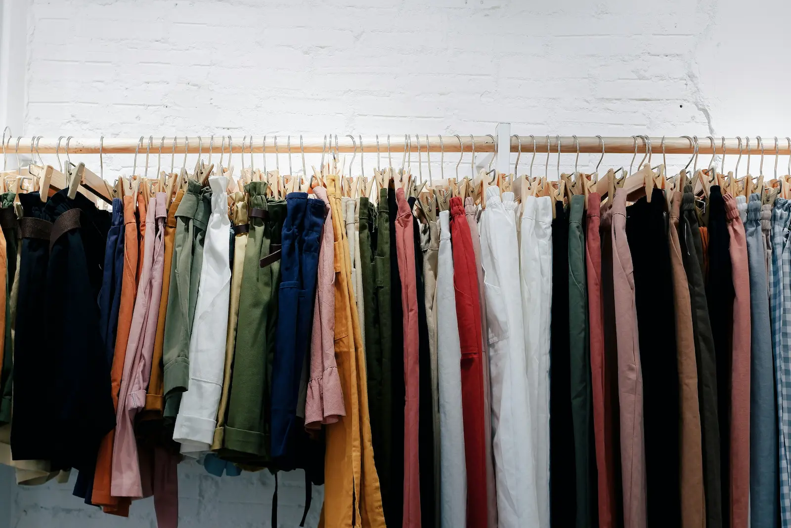 Colourful clothes hanging on a wooden rail against a white brick wall.